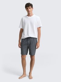 Ung mand står barfodet iført en simpel hvid t-shirt og marineblå rutede shorts foran en lys grå baggrund.