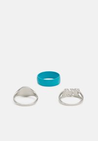 Três anéis sobre um fundo branco: uma faixa de silicone turquesa, um anel triangular em prata polida e um anel de prata com design recortado.