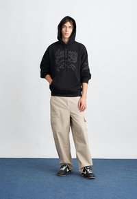 Svart huvtröja med grafiskt tryck, beige cargobyxor och svarta sneakers på en blå matta. Modellen står framför en enfärgad bakgrund.