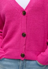 Helles pinkes Strickcardigan mit drei dunklen Knöpfen, getragen über hellblaue Jeans mit den Händen in den Taschen.