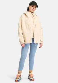 Beige waterdichte jas met een hoge kraag en ritszakken, gecombineerd met blauwe leggings en multikleurige sportschoenen.