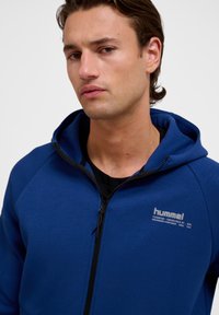 Modrá mikina na zips vyrobená z mäkkej látky s kapucňou, s logom na hrudi a čiernym zipsom. Jednoduchý dizajn s raglánovými rukávmi.