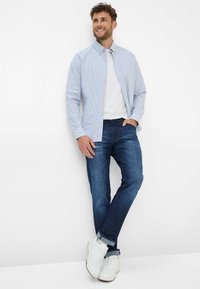 Lichtblauw gestreepte button-down shirt over een wit T-shirt, gecombineerd met donkerblauwe jeans en witte sneakers, staand tegen een witte muur.