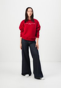 Červená mikina s logem "TOMMY HILFIGER EST. 1985", spárovaná s širokými tmavomodrými džíny. Bílé tenisky dokončují celkový vzhled.