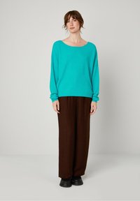 Turquoise oversized trui met lange mouwen, gecombineerd met bruine wijde broek. De trui heeft een ronde halslijn en een zachte textuur. Zwarte schoenen.