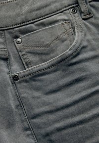 Grå denimjeans med en frontlomme, sort hardware og tonet syning. Stoffet viser en subtil struktur og let fading.