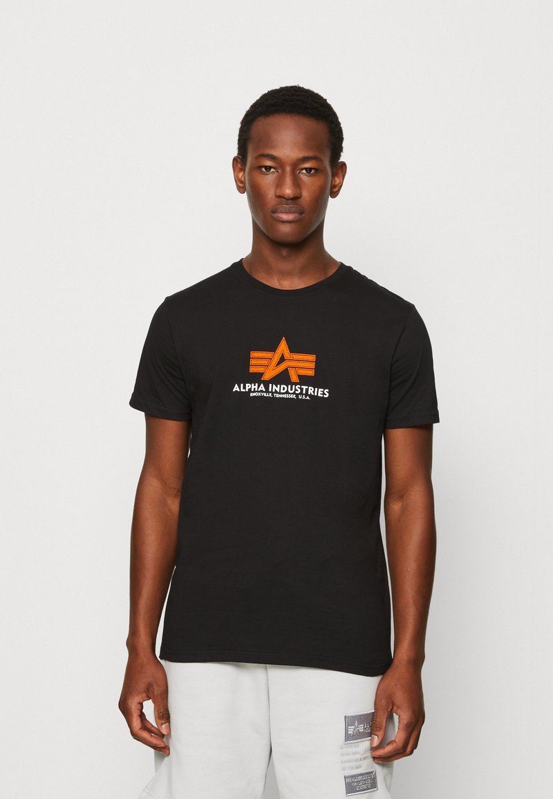 Juodasis medvilnės marškinėlis su oranžiniu ir baltu grafiniu logotipu ant krūtinės. Klasikinis apvalus kaklas ir trumpi rankovės. Kasdienis siluetas.