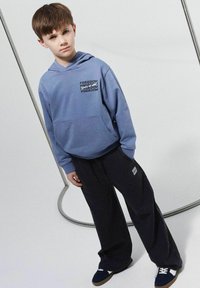 Helehelesin sinine kapuutsiga särk, millel on esiküljel tasku ja must graafiline tekst, koos avarate tumesiniste pükste ja tumedate kingadega. Minimalistlik disain.