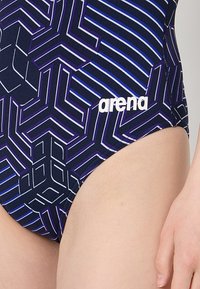 Maillot de bain avec un motif géométrique en bleu foncé, violet et blanc. Comprend le logo "arena" en blanc sur le côté. Texture lisse.