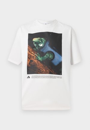 Valkoinen puuvillapaita, jossa on vihreä iguana-grafiikka mustalla taustalla. Alla oleva teksti tarjoaa tietoa designista.