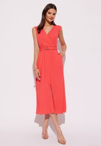 Korale, bezrękawna sukienka w stylu kopertowym z dekoltem w kształcie litery V, z paskiem w talii z okrągłą klamrą, zwiewna sylwetka oraz boczne kieszenie. Przezroczyste sandały.