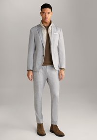 Lichtgrijs op maat gemaakt pak van textuurstof, gecombineerd met een bruine gebreide trui en een beige colbertshirt. Bruine suède enkellaarsjes.