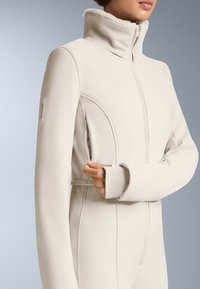 Jasnoszara, teksturowana odzież wierzchnia z wysokim kołnierzem, zamek błyskawiczny z przodu i dyskretną kieszenią boczną. Posiada logo RECCO na rękawie.