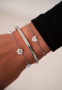 Pulseras de plata en una muñeca: una pulsera plana, una cadena con un charm de corazón y una con un diseño floral que presenta pequeños acentos de perlas.