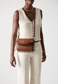 Smeđi kožni crossbody torba s pletenim detaljima, zlatnim metalnim naglascima i podesivim remenom, nosi je osoba u kremastom puloveru i svijetlim hlačama.