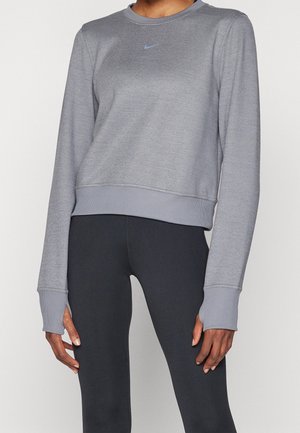 Osoba ubrana w jasnoszarą, długorękawową bluzę Nike z otworami na kciuki oraz czarne dopasowane legginsy na jednolitym tle.