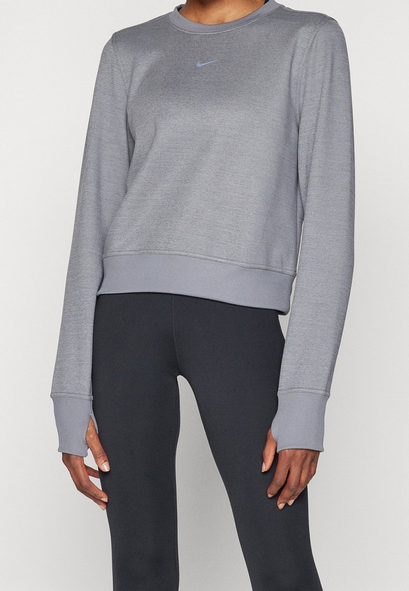 Osoba ubrana w jasnoszarą, długorękawową bluzę Nike z otworami na kciuki oraz czarne dopasowane legginsy na jednolitym tle.