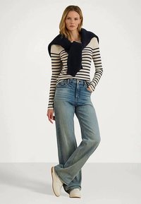 Tânără stând în picioare cu mâna în buzunar, purtând un tricou cu dungi pe mânecă lungă, blugi evazați, pantofi albi și un pulover întunecat aruncat peste umeri.