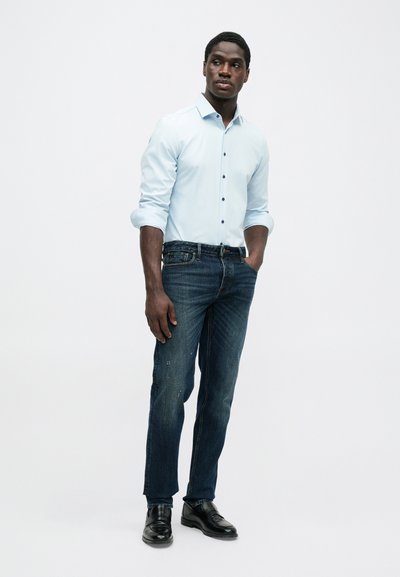 Ανοιχτό μπλε πουκάμισο με κουμπιά, σκούρο μπλε τζιν και μαύρα παπούτσια. Το πουκάμισο διαθέτει γιακά και γυρισμένα μανίκια, ενώ το τζιν έχει στενή εφαρμογή.