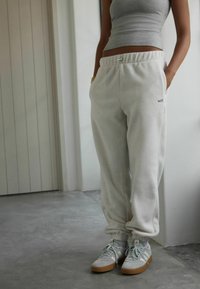 Inimene kannab heledat halli dressipükse, millel on kiri "self.", halli värvi T-särki ja halli Adidas'i spordijalatsit, millel on pähklikoore värvi tallad, seismas siseruumides.