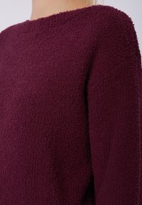 Pull bordeaux texturé avec un matériau doux et duveteux, un col large et des manches longues. Gros plan sur le détail de la texture en boucles.