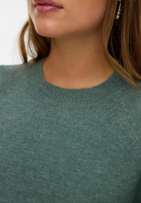 Vrouw in een groene gebreide trui met een geribde kraag en een hangende pareloorbel, die haar nek en een deel van haar gezicht laat zien.