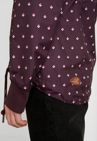 Lila jacka med vit och röd diamantmönster, elastiska ärmslut och en strukturerad beige logotyp på den nedre fållen. Tyget verkar vara slätt.