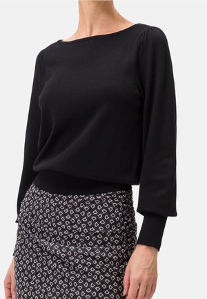 Haut noir à manches longues avec un col large, en tissu doux, et des accents froncés aux épaules, associé à une jupe à motifs avec des courbes.