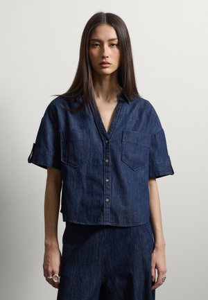 Joven mujer con cabello largo y oscuro lleva una camisa vaquera oscura de manga corta con botones y pantalones a juego, de pie sobre un fondo claro.