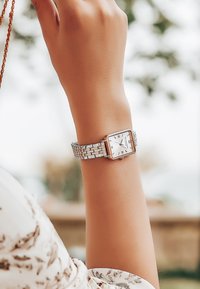 Reloj rectangular de tono plateado con una esfera blanca, números romanos y una pulsera de metal texturizada, mostrado en una muñeca.