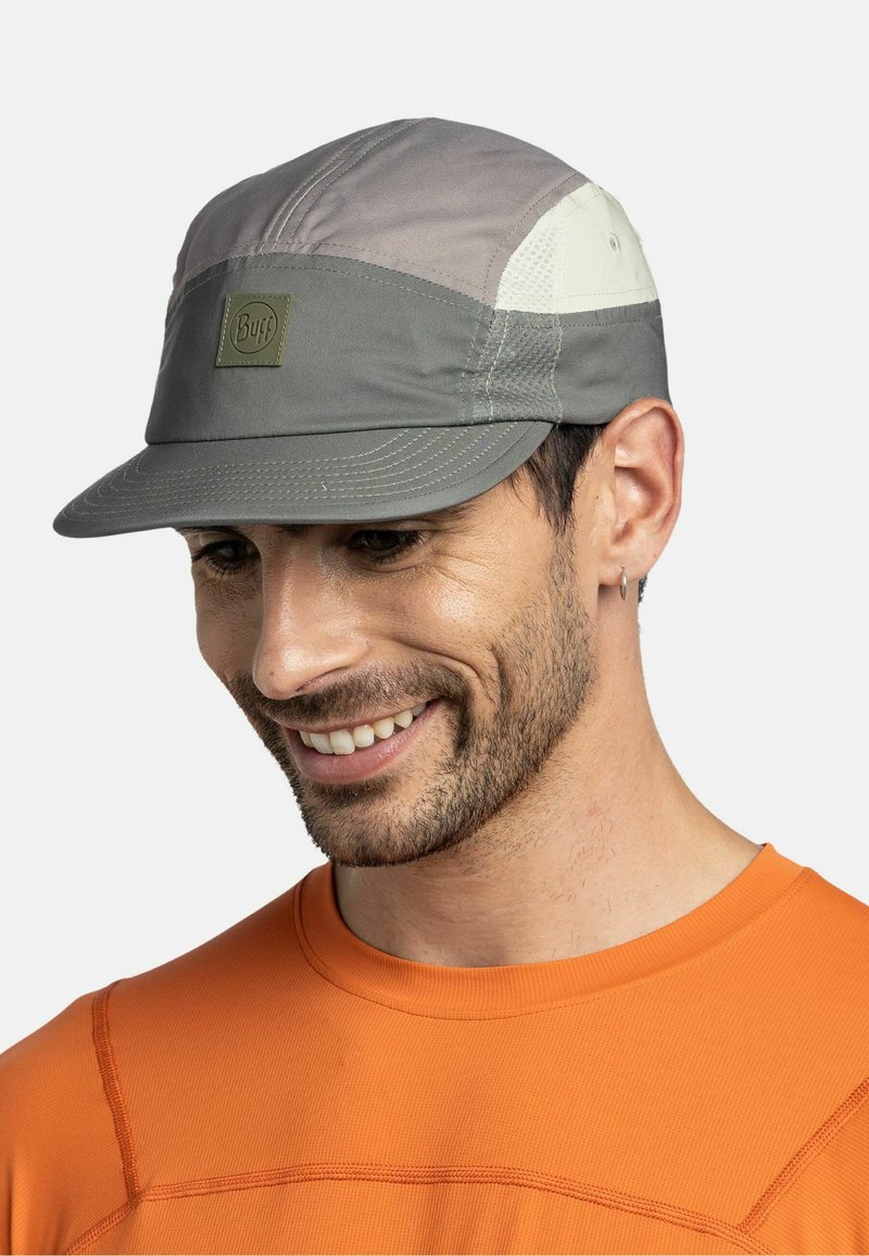 Man draagt een grijs en beige pet met het "Buff"-logo, een oranje shirt, glimlachend en naar beneden kijkend, tegen een effen lichte achtergrond.
