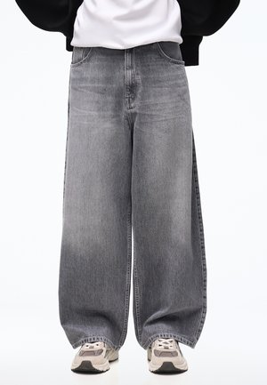 Personne portant un jean gris délavé à jambes larges et des baskets blanches avec des accents beige et noir, debout sur un fond uni.