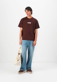 Young man wearing a brown Tommy t-shirt, light blue wide-leg jeans, brown sneakers, holding a large beige tote bag against a plain white background.