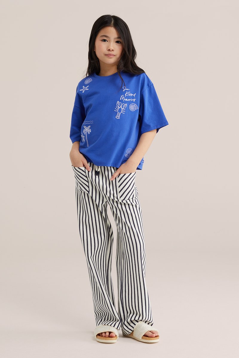 Jeune fille debout, les mains dans les poches, portant une chemise bleue à manches courtes avec des motifs marins et un pantalon blanc à rayures verticales noires.