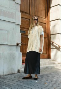 Gilet en tricot à coupe ample, couleur crème et fermeture par bouton, superposé à une chemise blanche et une jupe en satin noire, assorti à des chaussures plates noires.