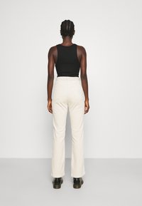 Top preto sem mangas com design ajustado, combinado com calças beiges claras e em formato boca de sino, apresentando bolsos traseiros e um tecido texturizado. Botas pretas até ao tornozelo.