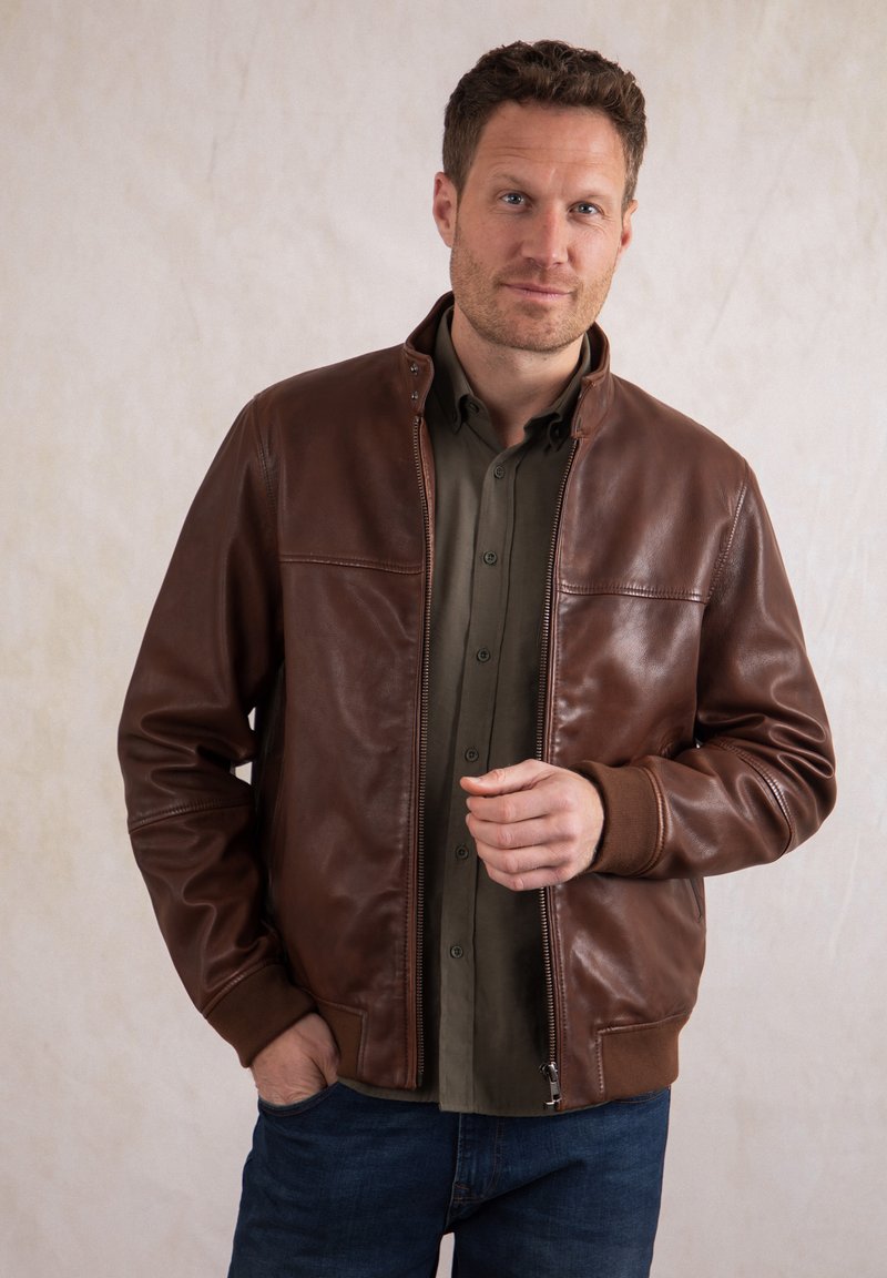 Man wearing brown leather jacket over olive shirt and blue jeans, standing with one hand in pocket against plain background.