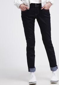 Osoba v tmavých slim-fit rifle s rolled-up nohavicemi, bílých teniskách a bílé bundě s rukama v kapsách na jednobarevném pozadí.