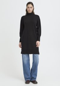 Nő fekete, hosszú ujjú garbóruhát visel kék farmerrel és barna cipővel, fehér egyszínű háttér előtt áll.