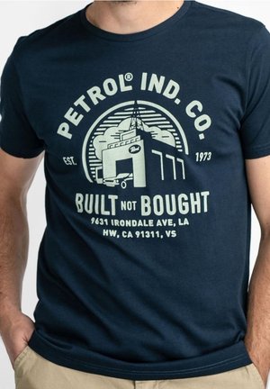 Muž nosící námořnicky modré tričko s logem Petrol Ind. Co. a textem „Built Not Bought“ a grafikou průmyslové budovy na přední straně.