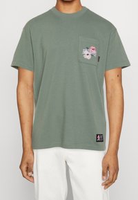 Green cotton t-shirt with a crew neck, pocket on the left chest featuring embroidery, and a logo patch at the bottom left hem.