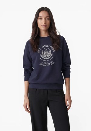 Kvinde med langt mørkt hår iført en marineblå sweatshirt med hvid Neo Heritage Berlin tekst og grafik, kombineret med sorte bukser.