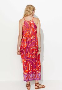 Robe maxi paisley rouge et violet avec bretelles spaghetti, dos découpé et ourlet détaillé ; portée avec des sandales brunes à lanières.