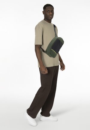 Man wearing beige t-shirt, dark brown pants, white sneakers, and green and black crossbody bag standing against white background.