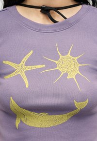 Μοβ πουκάμισο με κίτρινα τυπωμένα θαλάσσια πλάσματα: αστερίας, σχήμα σαν ήλιος και φάλαινα, φορεμένο από άτομο με μαύρο κορδόνι στο λαιμό.