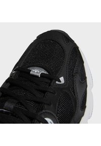 Shoe atlético preto com parte superior em malha e sintético, biqueira reforçada, detalhes circulares e atacadores planos. Sola branca com piso texturizado.