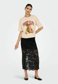 Camiseta de algodón beige con un gráfico de oso de peluche y texto "Teddy Club", combinada con una falda larga negra de encaje con patrones florales.