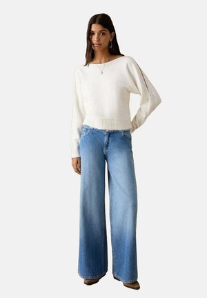 Pull blanc en maille avec ourlet côtelé, associé à un jean large bleu clair. Le jean présente un effet délavé et un design classique à cinq poches.