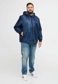 Svetlo modra kapuca jakna temno modre barve z bleščečo zunanjostjo, zadrgo in črnimi manšeti. Model nosi svetlo modre kavbojke in bele superge.