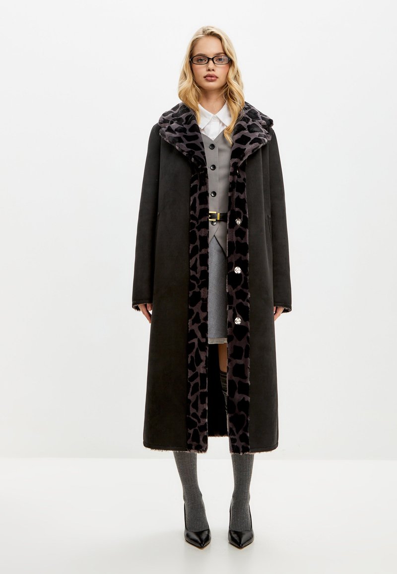 Lange zwarte jas met een voering van luipaardprintbont, voorzien van grote knopen, zijzakken en een oversized kraag. Gestyled met een grijze jurk en grijze sokken.